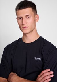 Fekete, rövid ujjú pamut póló, kerek nyakkivágással és logóval a bal mellen. A modellen keresztbe tett karokkal, semleges háttér előtt.