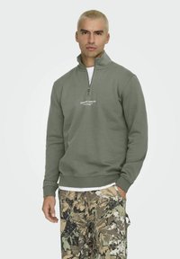 Pulover verde măsliniu cu fermoar la jumătate, cu mansete cu model în valuri, având un logo alb, purtat peste un tricou alb, asortat cu pantaloni în stil camuflaj.