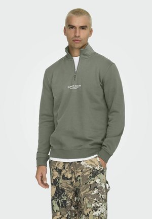 Olive green quarter-zip sweatshirt with ribbed cuffs, featuring a white logo and worn over a white t-shirt, paired with camo pants.