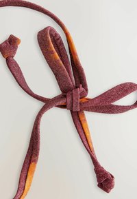 Tumman violetin värinen tekstiilitaskuliina, jossa on oransseja yksityiskohtia, ja joka esittelee solmittua muotoilua sekä kiiltävän, tekstuurin omaavan pinnan.