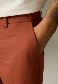 Pantaloni de culoare ruginită, cu o textură netedă și buzunare tradiționale. Au un front plat și un talie ajustată fără accesorii vizibile.