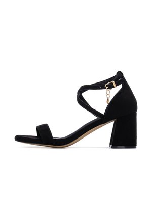 Sandalia de tacón de ante negro con tacón cuadrado y diseño de tiras cruzadas. Presenta una pequeña hebilla y detalle de cadena en la correa del tobillo. Textura suave.