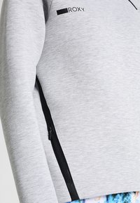 Sweat-shirt gris à texture lisse, avec une fermeture éclair noire sur le côté et un logo noir sur la poitrine.