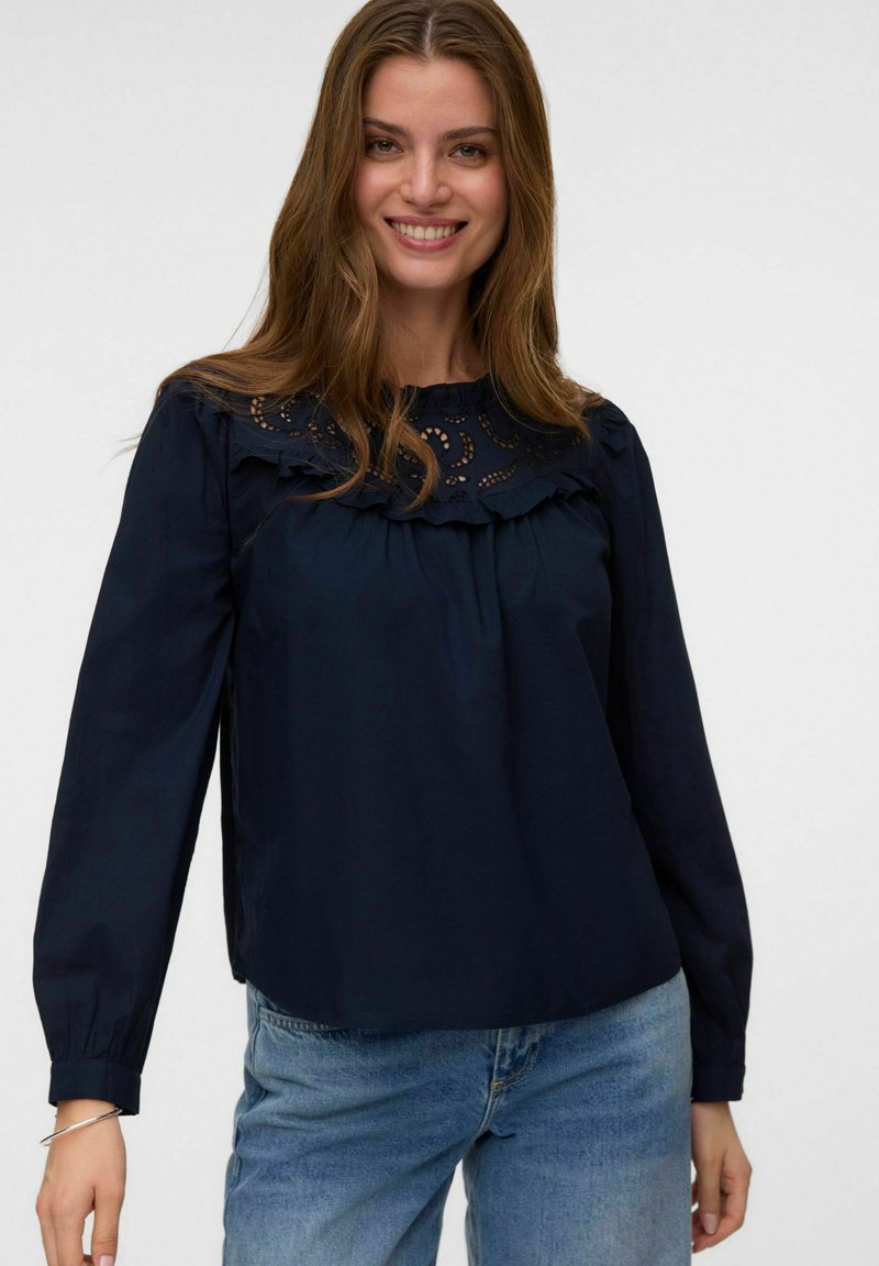 Jeune femme souriante, portant un chemisier bleu marine à manches longues avec des détails en dentelle et un jean bleu, debout devant un fond uni.