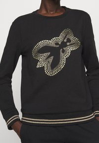 Sudadera negra con diseño de tachuelas metálicas doradas en la parte frontal, con cuello redondo acanalado y ribete a rayas en los puños y el dobladillo.