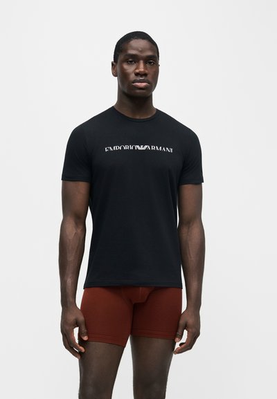 Άνδρας που φοράει μαύρο t-shirt Emporio Armani και σκουρόχρωμα, εφαρμοστά σορτς, στέκεται μπροστά σε απλό λευκό φόντο.