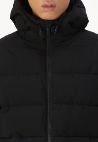 Sort puffjakke med høj krave, lynlås og snoretræk i hætten. Stoffet ser glat ud med en polstret tekstur.