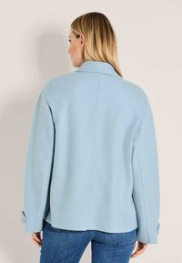 Eine Frau mit blonden Haaren, die eine hellblaue Wolljacke undblaue Jeans trägt, von hinten vor einem einfarbigen Hintergrund gesehen.