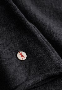 Donkergrijs stof met een zachte textuur, voorzien van een ronde metalen badge met een rode hondenopslag en een bruine draadlus.