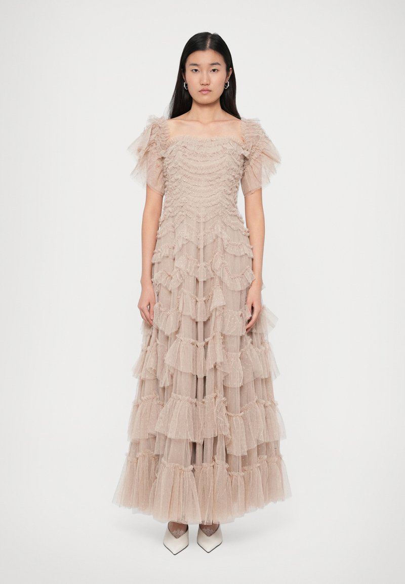 Pehmeas beežis tüllkleidis, millel on volangilised kihid, avatud õlgadega puffvarrukad ja voolav siluett, mida täiendab terav tipuga valge kingad.