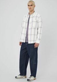 Chemise à carreaux blanche et violette sur un t-shirt gris, associée à un pantalon en denim bleu ample et des chaussures de sport blanches avec des accents noirs.