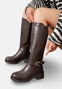 Bottes marron jusqu'au genou en cuir lisse avec un embout rond, accent de boucle et semelle texturée. La tige est droite et sans motifs.