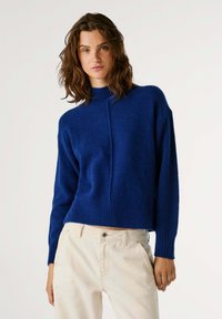 Mėlynas megztinis su aukštu kaklu, gipiūro apvadu ir rankogaliais, pasižymintis laisvu siluetu ir tekstūruotu audiniu. Derinamas su šviesių atspalvių kelnėmis.