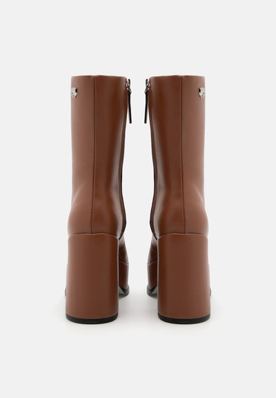 Bottines en cuir brun avec un talon bloc haut, fermeture éclair et accents métalliques en haut. Texture lisse avec un design épuré.