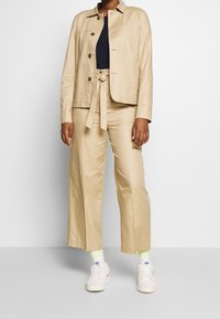 Giacca in lino beige con quattro bottoni e colletto, abbinata a pantaloni beige a gamba larga. Sneakers bianche con calzini a dettagli neon.