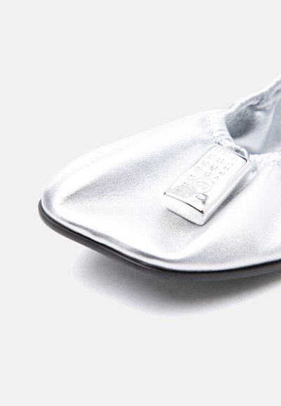 Κοντινό πλάνο λαμπερού ασημί μπαλαρίνα παπουτσιού με μαύρη σόλα και μεταλλική ετικέτα που εμφανίζει αριθμητικό σχέδιο στην περιοχή των δακτύλων.