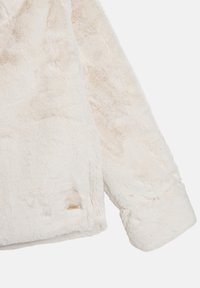 Giacca in pelliccia sintetica avorio con una textura morbida, caratterizzata da un orlo curvo e maniche arrotolate. Include un piccolo dettaglio del logo dorato.