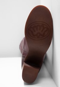 Burgundy läderstövel med en texturerad gummisula, som har en cirkulär logotyp och horisontella spår för grepp.