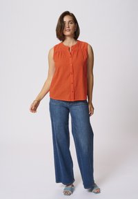 Haut sans manches orange à boutons avec des plis, associé à un jean en denim bleu à jambe large et des sandales bleues effilochées. Design simple et style décontracté.