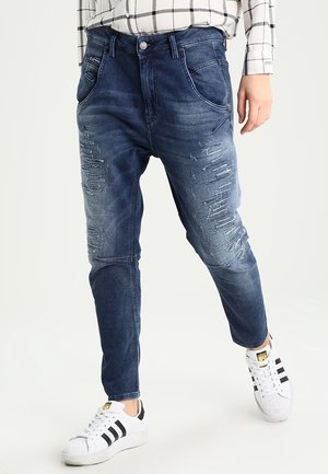 Osoba w niebieskich, znoszonych dżinsach z denimu połączonych z białymi sneakersami Adidas z czarnymi paskami, stojąca na białym tle.