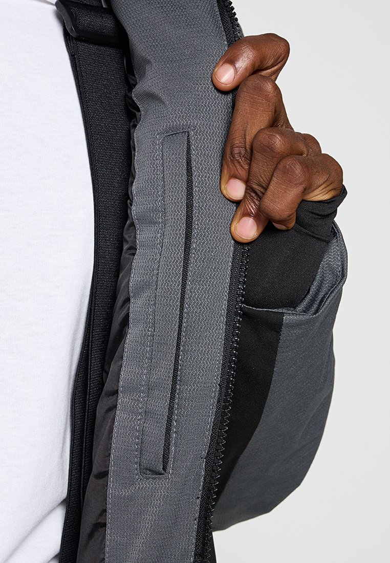 Mano de piel oscura sujetando la manga de una chaqueta gris y negra sin abotonar sobre una camisa blanca, resaltando el bolsillo interior de la chaqueta y los detalles de la cremallera.