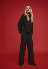 Czarna, dopasowana marynarka z dwoma przednimi kieszeniami, połączona z luźnymi czarnymi spodniami. Modelka nosi czarne buty na szpic z ozdobnymi elementami.