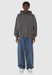 Grå hettegenser med kengurulomme, med brodert tekst. Kombinert med løse blå jeans og hvite joggesko.