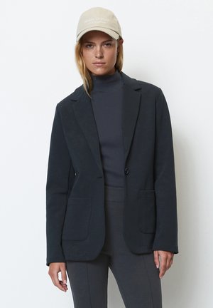 Dunkelmarineblaues Blazer mit maßgeschneidertem Schnitt, strukturiertem Stoff, zwei vorderen Taschen und einem einzelnen Knopfverschluss, kombiniert mit einem passenden Rollkragenpullover.