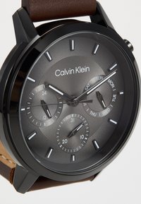 Montre en acier inoxydable noir avec un cadran texturé gris, des index horaires blancs, trois sous-cadrans et un bracelet en cuir marron. Marque : Calvin Klein.