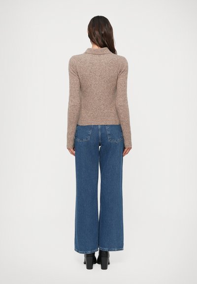 Mujer con pelo largo y oscuro, vistiendo un suéter beige de mangas largas, jeans anchos azul y botas de tacón negras, de pie contra un fondo liso.