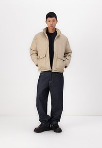 Beige dunjakke med høy krage, glidelås og frontlommer. Kombinert med mørkeblå baggy jeans og svarte klumpete sko.