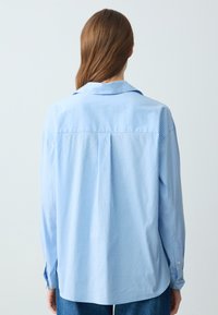 Lichtblauw langwerpig overhemd met verticale witte strepen, een kraag, knoopmanchetten en een plooi aan de achterkant voor extra detail.