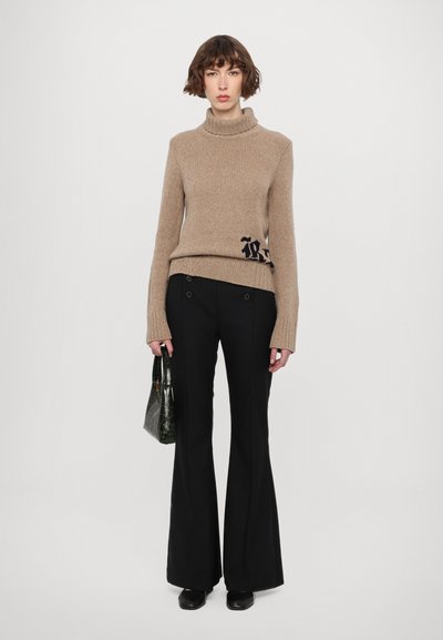 Beige gerippter Rollkragenpullover mit navyfarbener Logodetail, kombiniert mit schwarzen Schlaghosen. Das Model hält eine grüne strukturierte Handtasche.