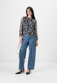 Modella in posa con un top blouson floreale e jeans a gamba larga, mentre tiene in mano una piccola borsa bianca, su uno sfondo bianco neutro.