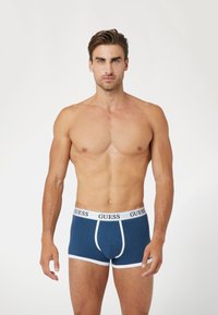 Boxers azul marino con cinturilla elástica blanca que presenta el logo "GUESS" y borde blanco. Tejido suave, diseño de longitud hasta mitad del muslo.