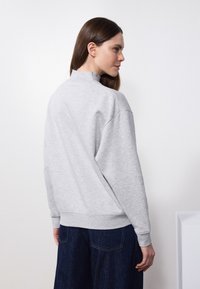 Sudadera de fleece gris con un alto cuello y hombros caídos, con puños y dobladillo acanalados. Combinada con pantalones de mezclilla azul oscuro.
