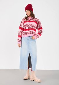 Γυναίκα με ροζ και κόκκινο καρό πουλόβερ, κόκκινο σκουφάκι, ανοιχτό μπλε τζιν φούστα με μπροστινή σχισμή, και μπεζ μποτάκια στον αστράγαλο, που στέκεται σε γκρίζο πάτωμα.