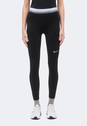 Žena nosi crne Nike tajice sa sivim pojasom i logotipom, sive čarape i bijele tenisice, stoji na svijetloj pozadini.