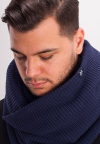 Knitted navy blue scarf with a chunky texture, wrapped around the neck, featuring a small logo tag on one side.