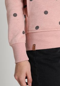 Felpa rosa con grandi pois grigi, polsini a costine e una piccola etichetta in pelle con il marchio nella parte inferiore dell'orlo, abbinata a tessuto nero.