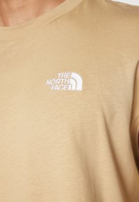 Tricou din bumbac bej, cu un logo brodat alb al "The North Face" pe partea stângă a pieptului. Design clasic cu gât rotund.