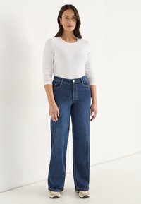 Kvinna klädd i en vit långärmad topp, högmidjade vida blå jeans, krämfärgade sneakers, som står mot en enkel ljus bakgrund.