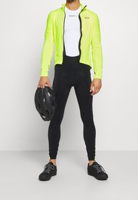 Giacca alta visibilità di colore verde neon con zip nera, indossata sopra un sottogiacca bianco. I pantaloni da ciclismo neri e le scarpe sportive nere completano l'outfit.