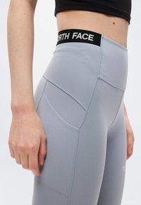 Κοντινό πλάνο ενός ατόμου που φοράει ανοιχτό γκρι κολάν υψηλής μέσης με μαύρη μέση που φέρει την ετικέτα "NORTH FACE" και πλαϊνή τσέπη.