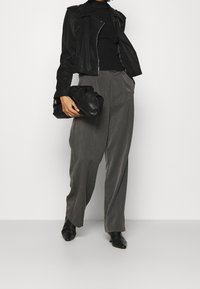 Giacca di pelle nera, dolcevita nera aderente, pantaloni grigi a gamba larga, scarpe nere, che tiene una pochette nera textures. Aspetto pulito e moderno.