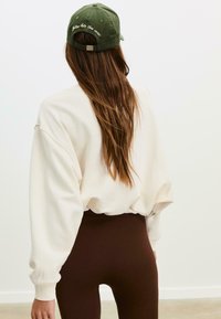 Felpa crema con spalle cadenti, abbinata a leggings marroni a vita alta. Un cappellino verde con testo ricamato completa l'outfit.
