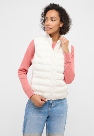 Frau mit lockigem Haar, die eine weiße Steppweste über einem rosa Pullover und blauen Jeans trägt und den Kragen der Weste richtet.