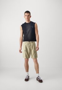 Top senza maniche nero con tasca con cerniera, pantaloni cargo beige con tasche laterali e sneakers marroni con strisce bianche. Design pulito e minimalista.
