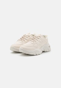 Beige synthetic sneakers with thick, zigzag-patterned soles, round toe design, and lace-up front; minimal detailing with subtle yellow accent.