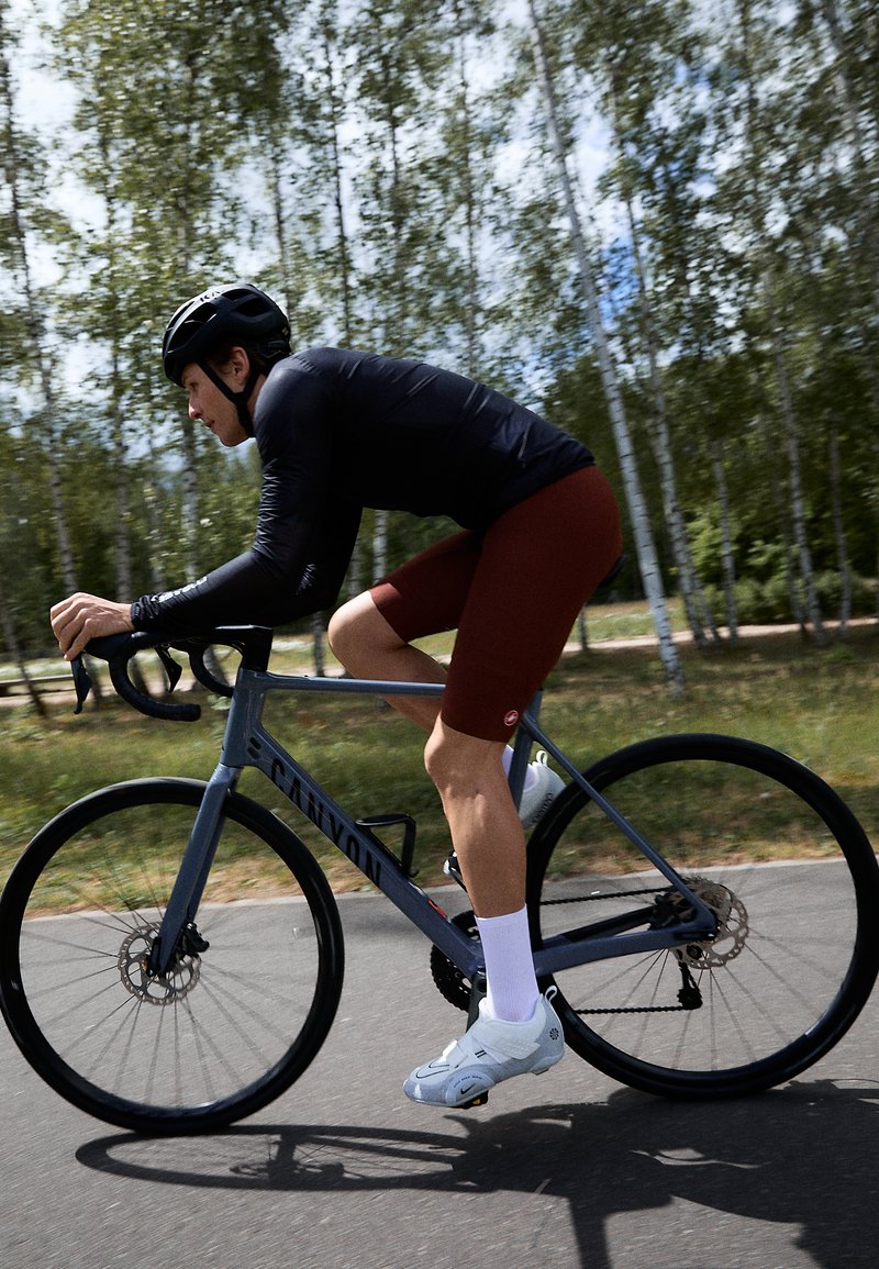 Grau Canyon Fahrrad mit schwarzen Rädern, Fahrer mit langem schwarzen Shirt und weinroten Shorts, weißen Socken und grauen Radsportschuhen auf asphaltierter Strecke.